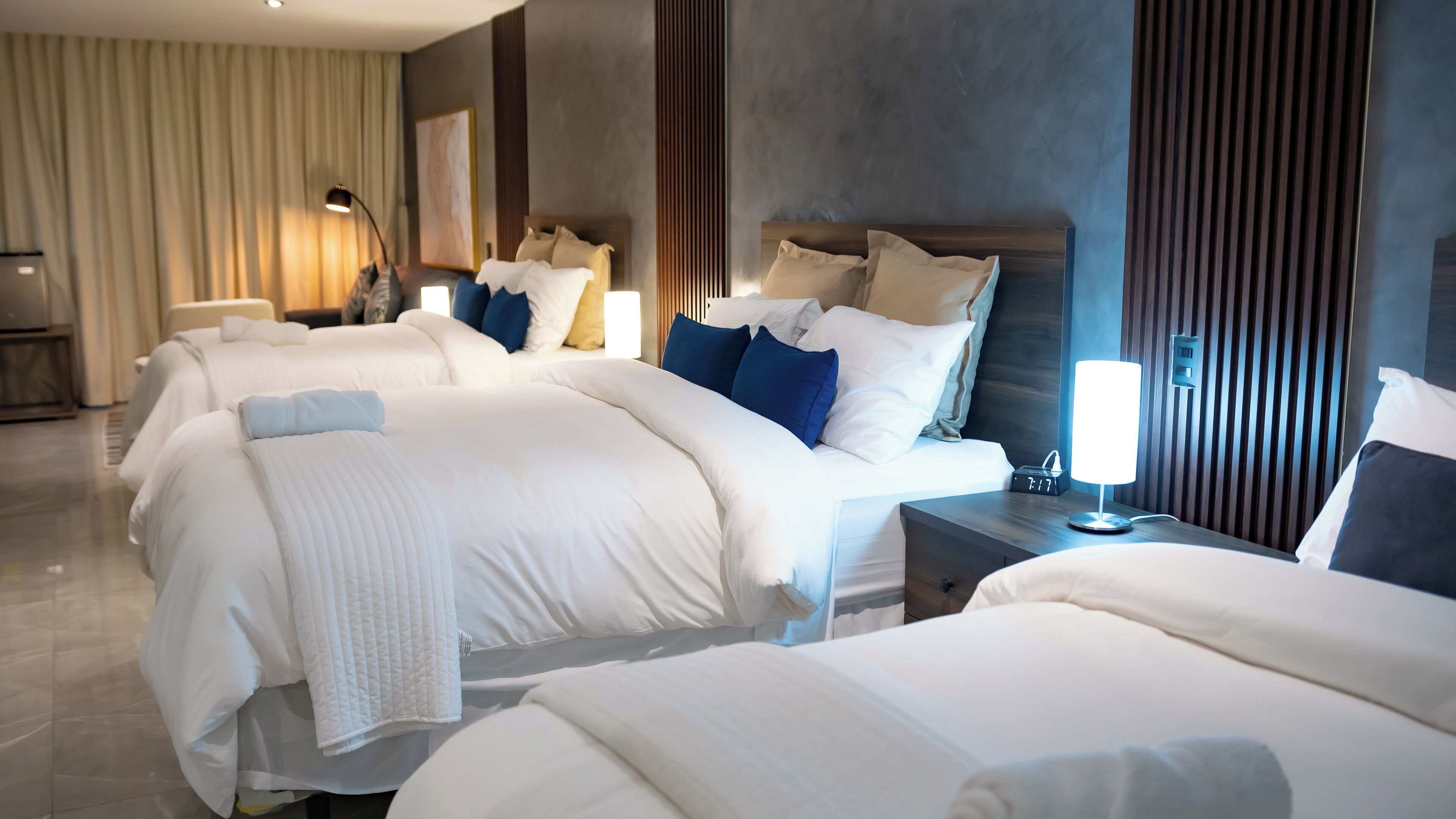 A hotel room with three beds, a nightstand with a lamp, and a large window with curtains.