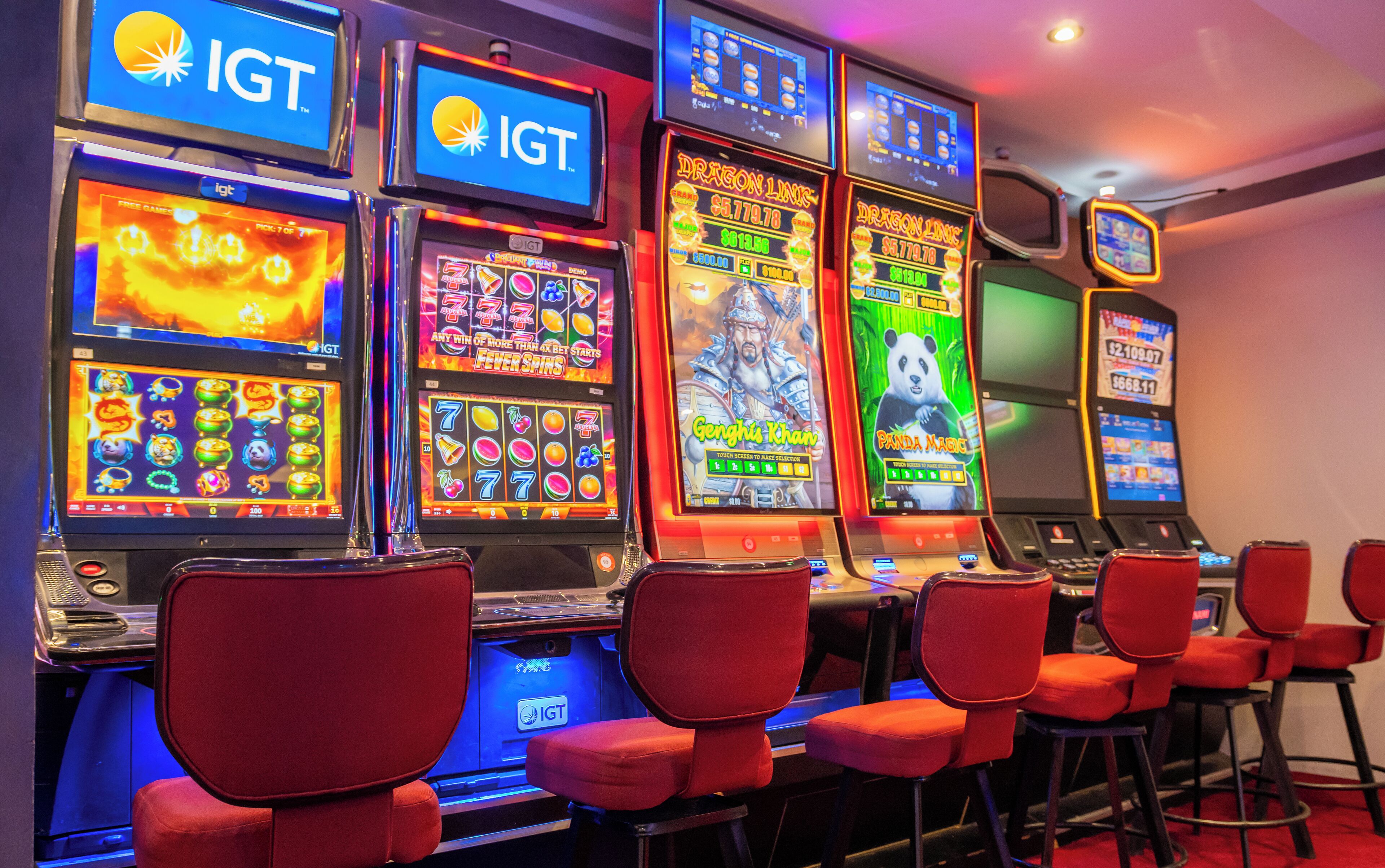 A row of slot machines with the IGT logo, red chairs in front, and a blue-lit ceiling.