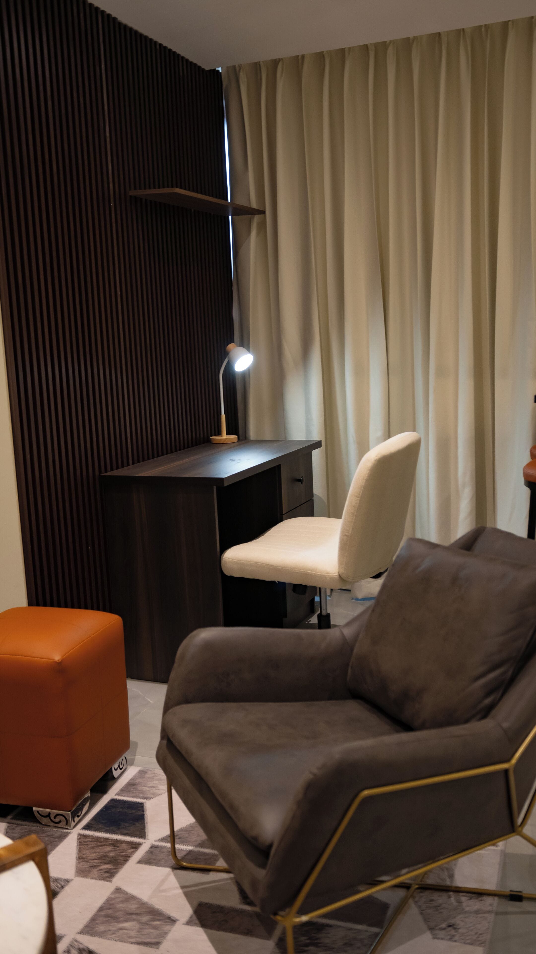 A modern room with a dark wooden desk, a white chair, an orange ottoman, and a lamp.
