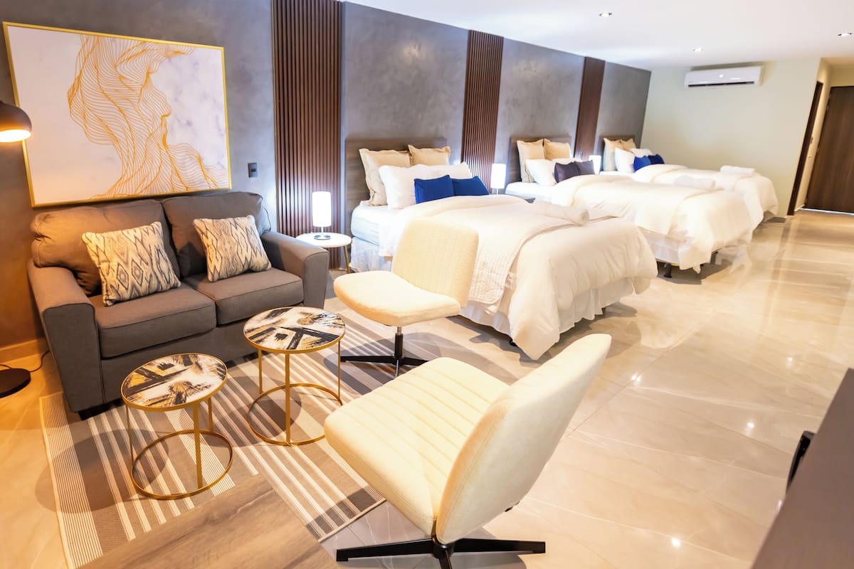 A modern hotel room with a sofa, armchair, two round tables, a large bed, and a wall-mounted artwork.