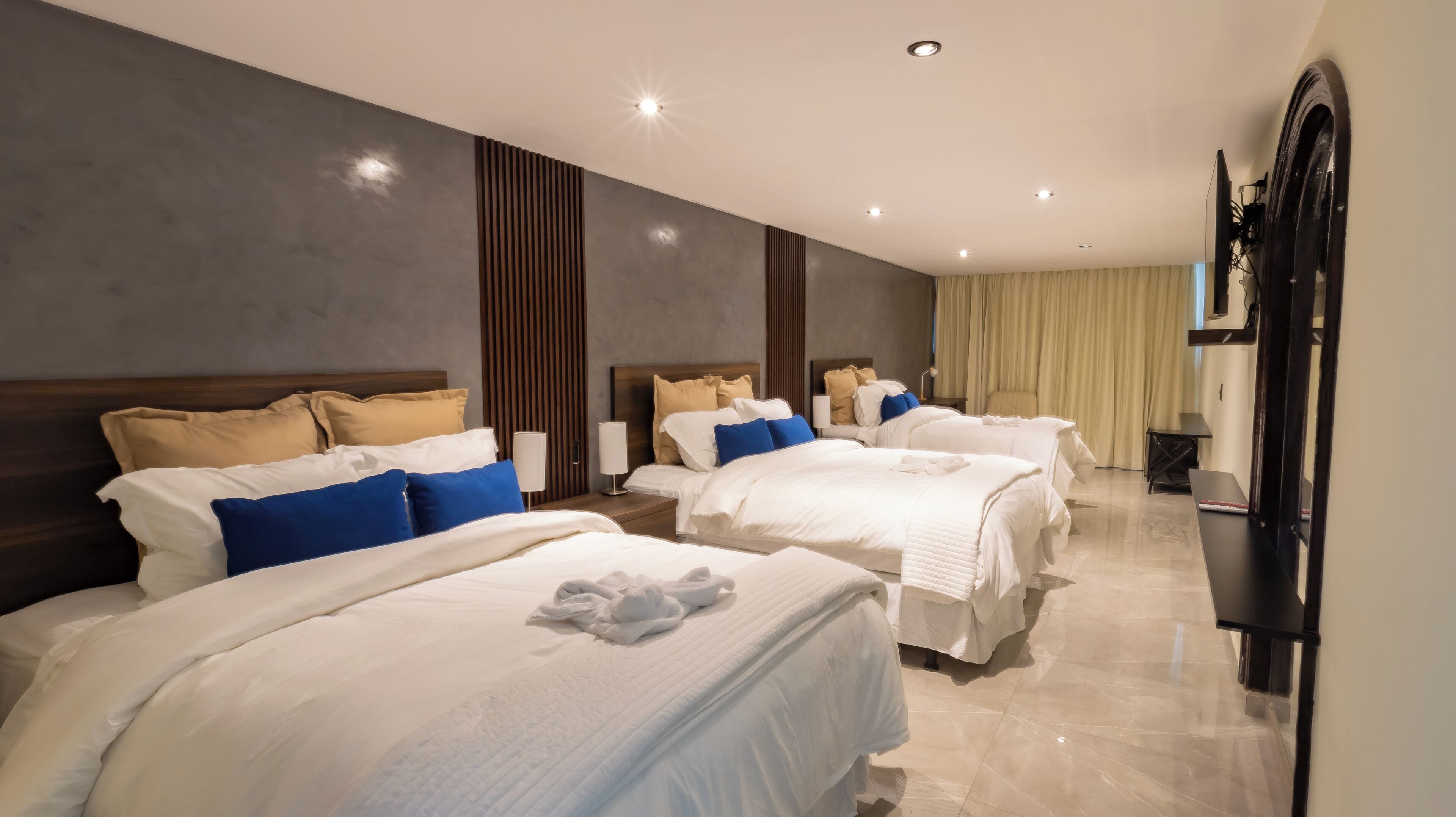 A hotel room with three beds, each with white bedding and blue pillows. The room has a modern design with a large mirror and a flat-screen TV mounted on the wall.