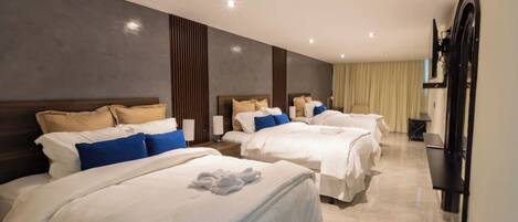 A hotel room with three beds, each with white bedding and blue pillows. The room has a modern design with a large mirror and a flat-screen TV mounted on the wall.