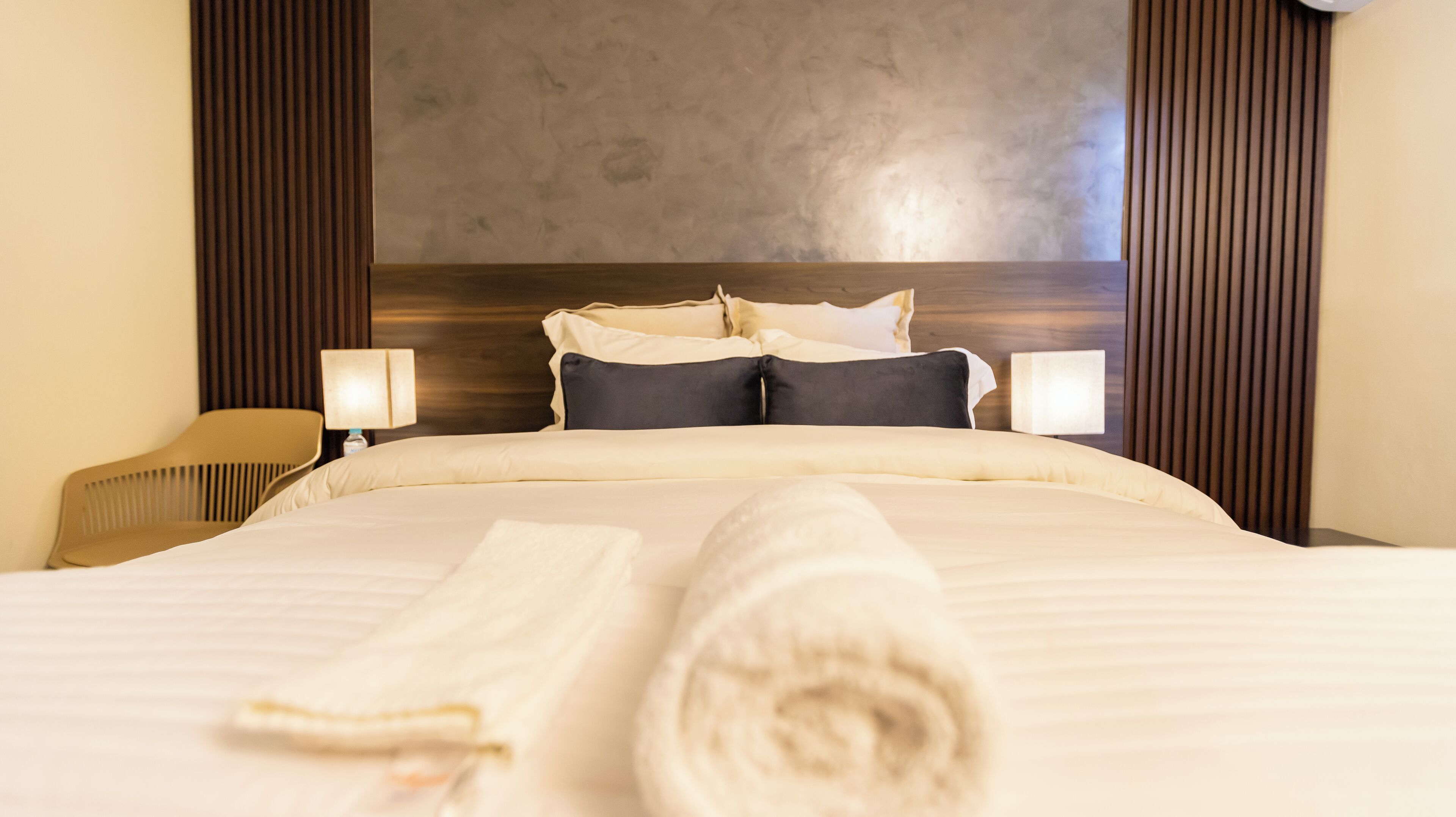 Habitación de hotel con una cama grande, dos lámparas de noche, un cabecero de madera y una toalla enrollada sobre la cama.