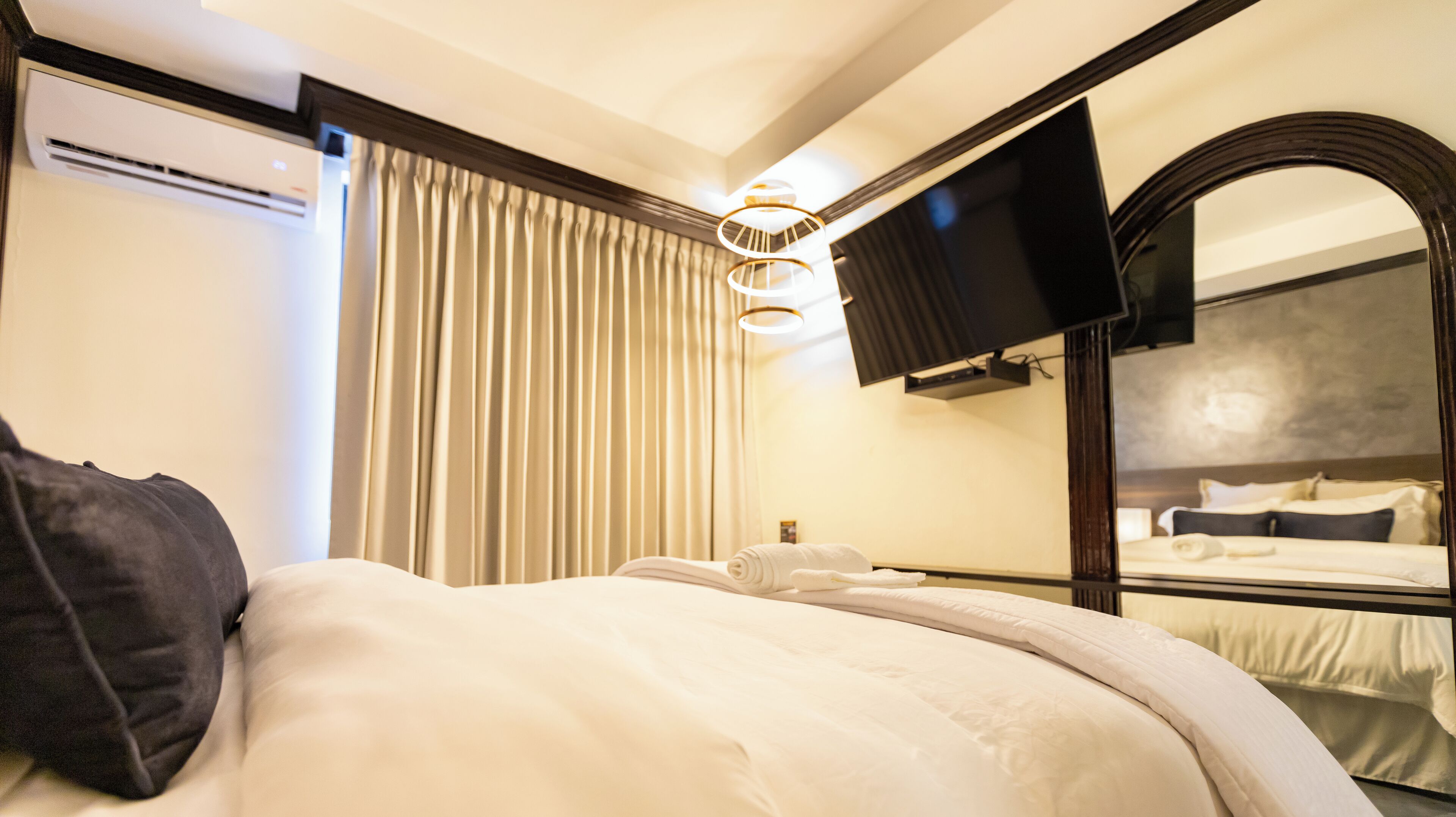 A hotel room with a bed, a large mirror, a flat-screen TV, and a ceiling light fixture.