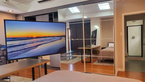A modern living room with a large flat-screen TV displaying a sunset view, a dining area, and a bedroom with a walk-in closet and shower. - Indoor and Outdoor Living (Los Angeles)
