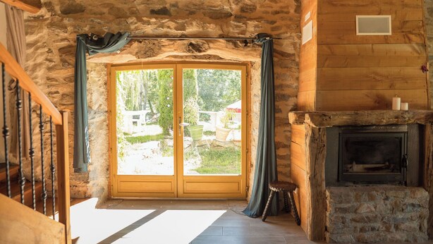 A log cabin interior with a stone wall, wooden beams, a fireplace, and a large window with curtains. - L'Ange : Entre monts et vallées House with billiard table and fiber optics (Retournac)