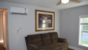 A living room with a brown couch, a ceiling fan, an air conditioner, and a framed picture on the wall.