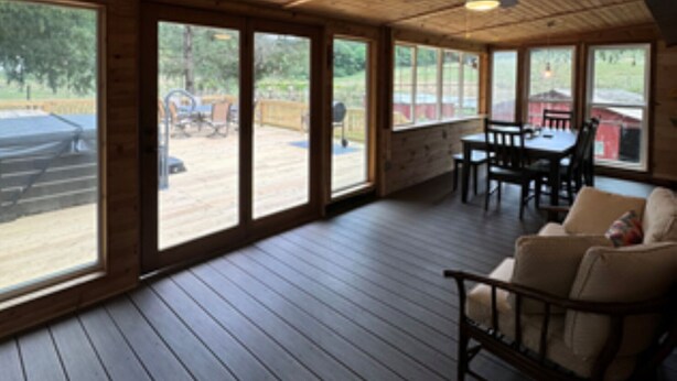 A covered outdoor area with wooden flooring, large windows, and a dining table with chairs.