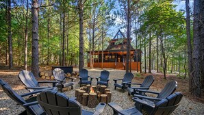 A cabin surrounded by tall trees, with a fire pit and Adirondack chairs in the foreground. - A Point in Time: Sleeps 12-16 | Fire Pit | Hot Tub (Broken Bow)