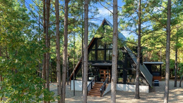 A modern cabin with a steep roof, surrounded by tall trees and gravel ground. - A Point in Time: Sleeps 12-16 | Fire Pit | Hot Tub (Broken Bow)