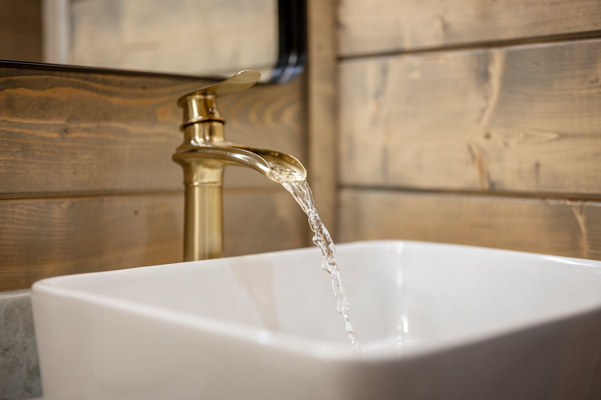 Un lavamanos blanco con agua corriendo de un grifo dorado sobre un pared de madera.