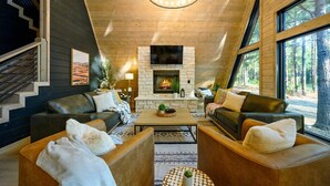 A modern living room with a fireplace, leather sofas, and large windows offering a forest view. - A Point in Time: Sleeps 12-16 | Fire Pit | Hot Tub (Broken Bow)
