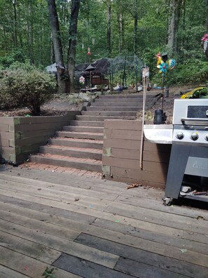 A wooden staircase leading up to a cabin in a forested area, with a grill and a bird feeder nearby.