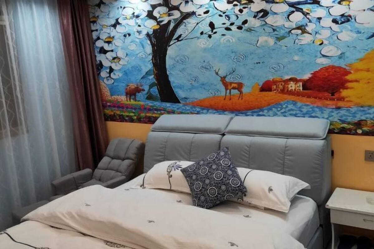 Habitación de hotel con cama, cabecera gris, sillón gris y un mural en la pared que muestra una escena natural con árboles, flores y animales.