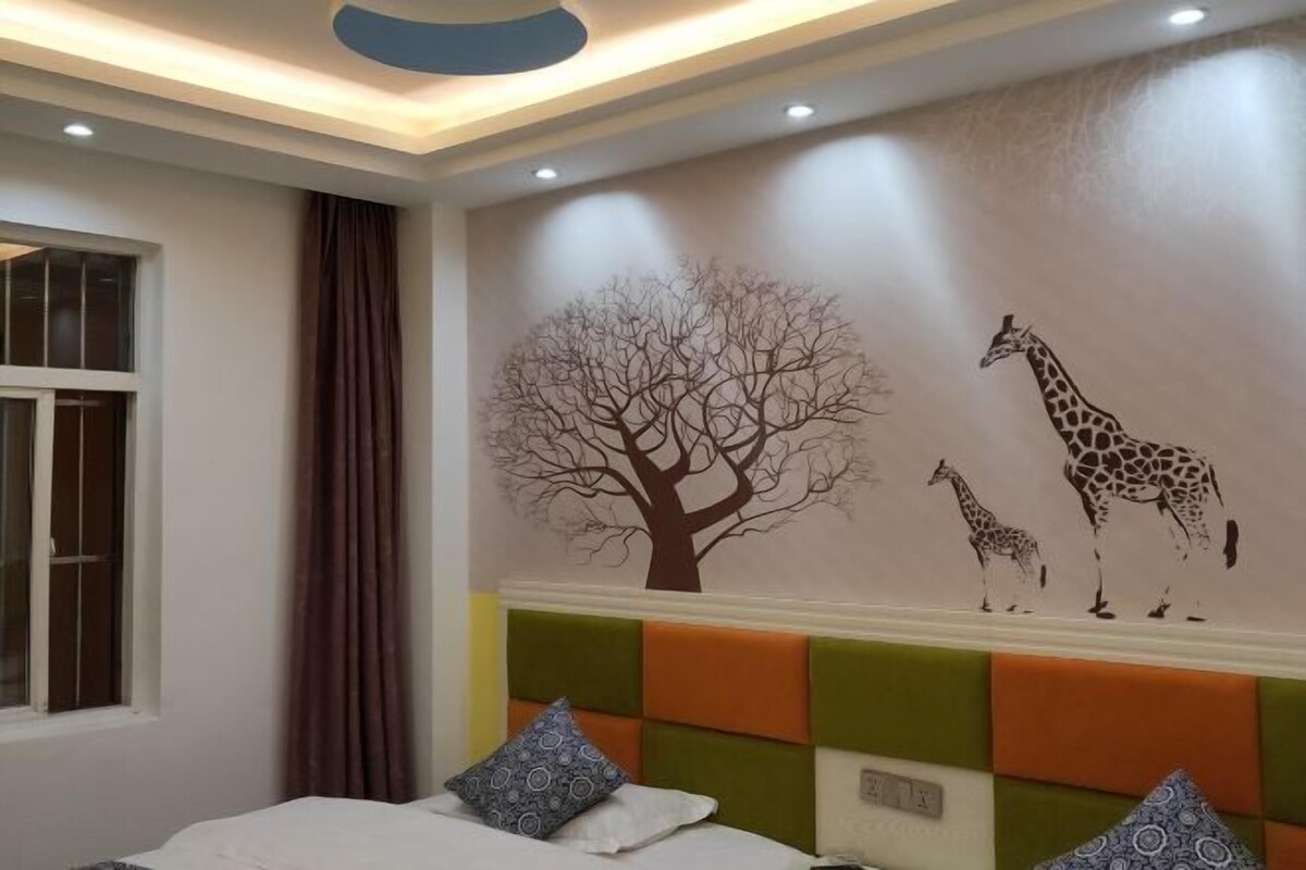 Un dormitori amb un llit, una finestra, una cortina, una paret amb un mural de girafa i arbre, i un capçal de llit amb un disseny colorit.