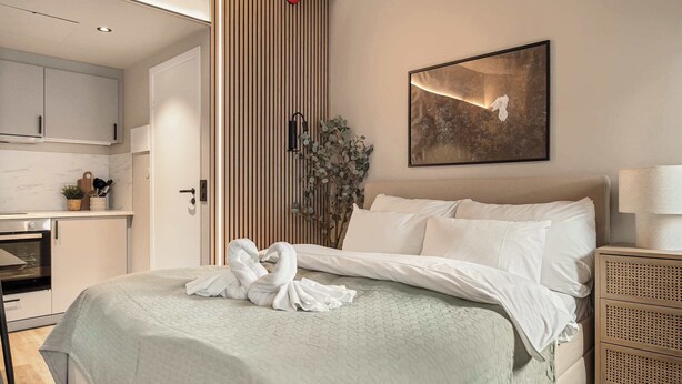 A hotel room with a bed, a bedside table, a lamp, a painting on the wall, and a kitchen area visible through an open door.