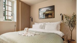 A neatly made bed with white linens, a large window with blinds, a wall-mounted picture, and a potted plant.