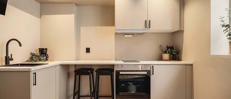 A compact kitchen with a built-in oven, two bar stools, a sink, and a window with a plant.