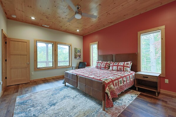 A bedroom with a wooden ceiling, a bed with a red and white quilt, a nightstand, and two windows with views of trees.