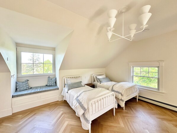 A bedroom with two white beds, a bench with cushions, a ceiling light fixture, and large windows.