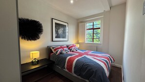 Ein Schlafzimmer mit einem Bett, einem gerahmten Bild an der Wand, einem Fenster mit Fensterläden und einer Lampe auf einem Nachttisch.