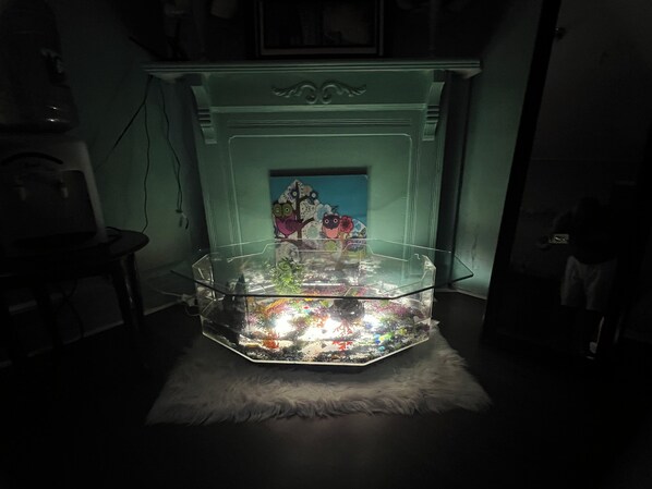 A room with a glass fish tank on a white furry rug, a green wall, and a white fireplace with a painting.