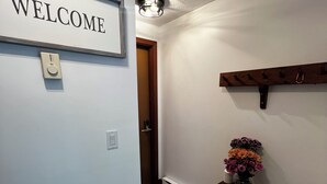 A hallway with a 'WELCOME' sign, a coat rack with a vase of flowers, and a thermostat on the wall. - 1BR apartment next to the Pleasant Mountain with waterfront access (Bridgton)