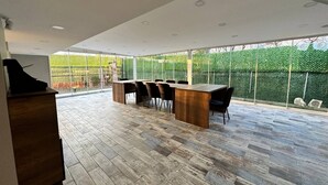 Amplio patio con mesa y sillas de madera, un banco incorporado y vistas a un seto verde a través de amplios ventanales.