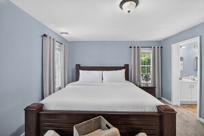 Une chambre à coucher avec un grand lit, une table de chevet, une fenêtre avec des rideaux, une porte donnant sur une salle de bain et un luminaire au plafond.