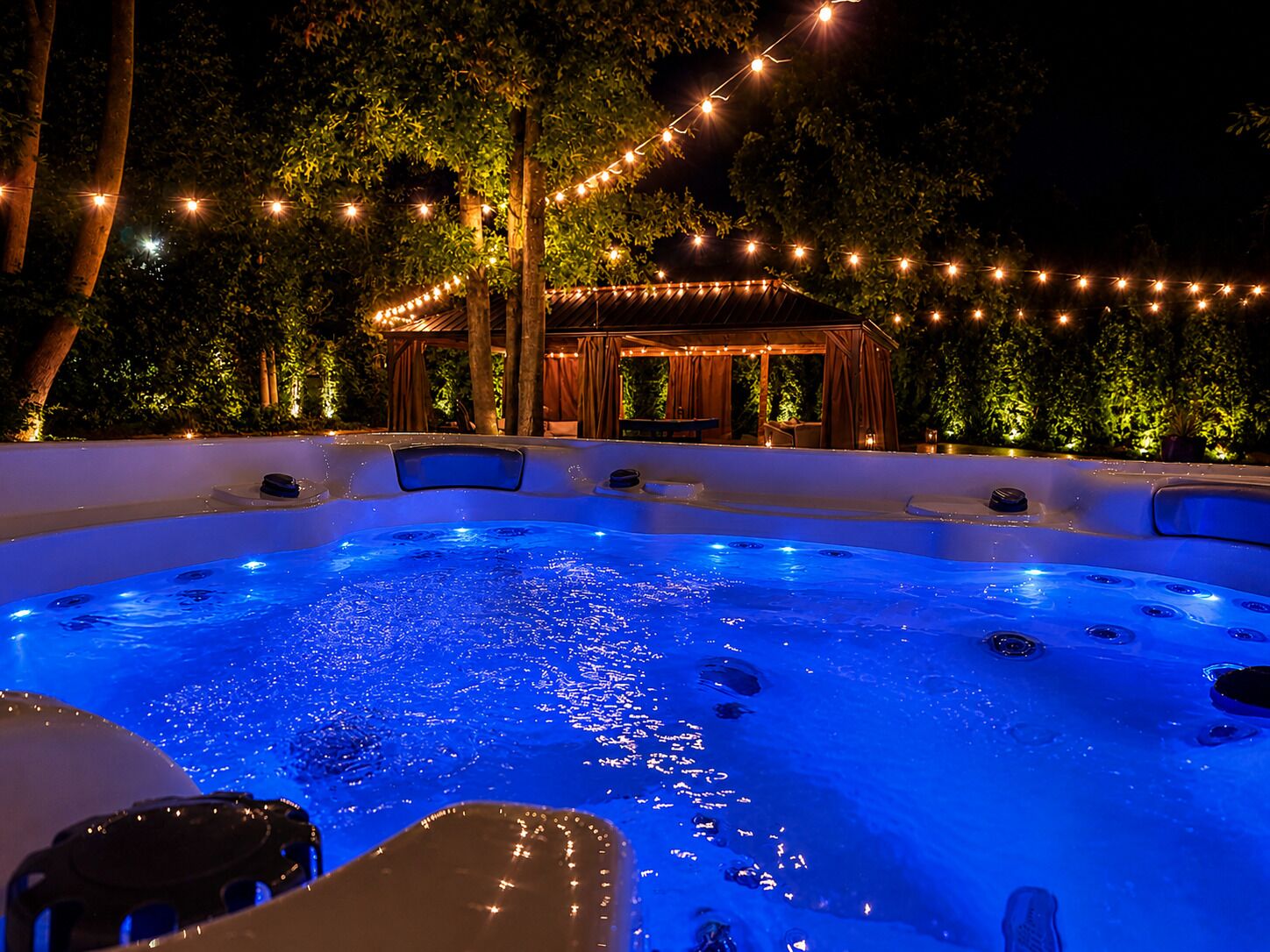 A hot tub with a blue light, surrounded by trees and string lights.