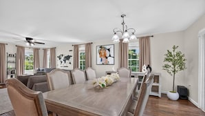 Une salle à manger avec une longue table, des chaises, un lustre, un ventilateur de plafond et une plante en pot.