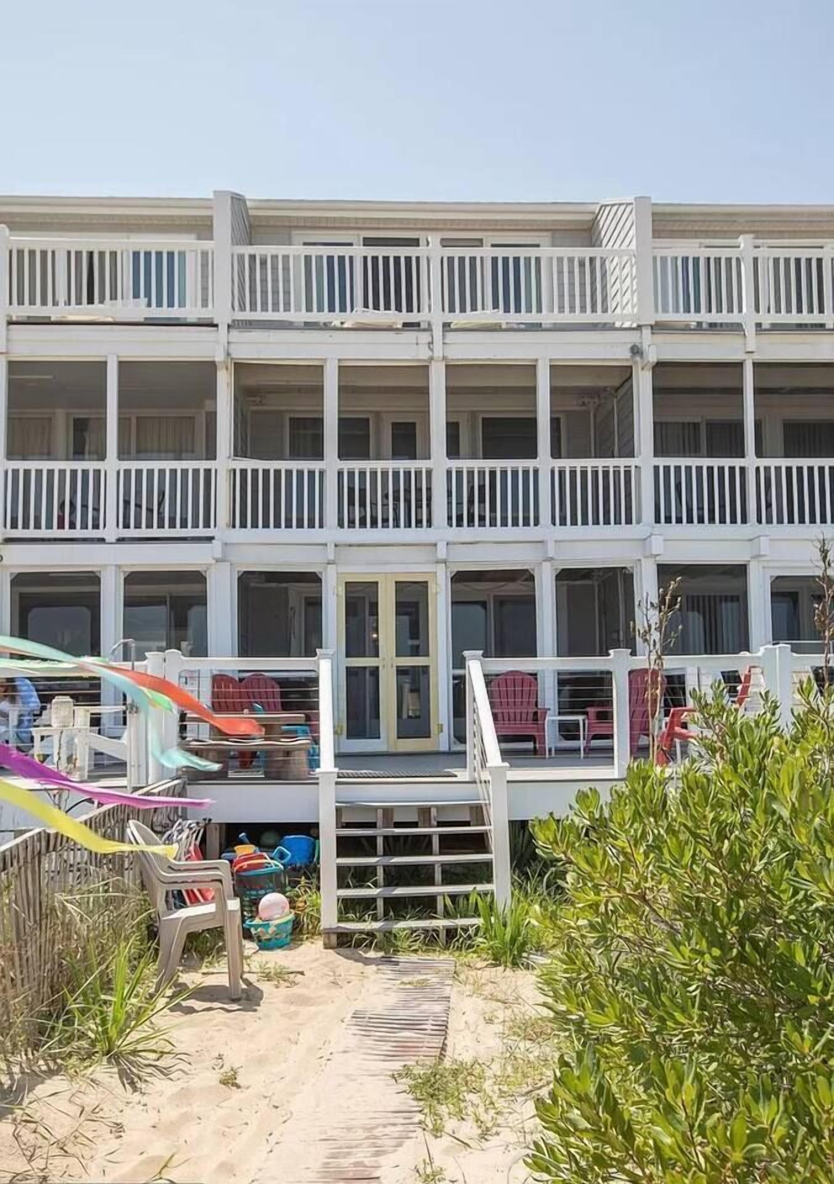 A multi-story building with balconies, a sandy ground, and some greenery.