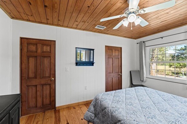 Un dormitorio con techo de madera, dos puertas, una cama con colcha estampada, una ventana con vistas y un ventilador de techo.