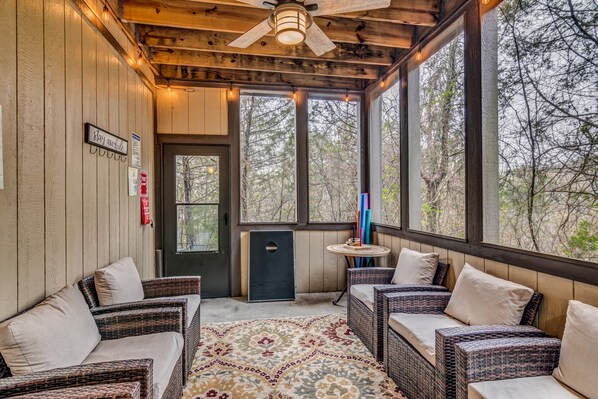 A covered porch with wicker furniture, a patterned rug, and a view of trees through large windows.