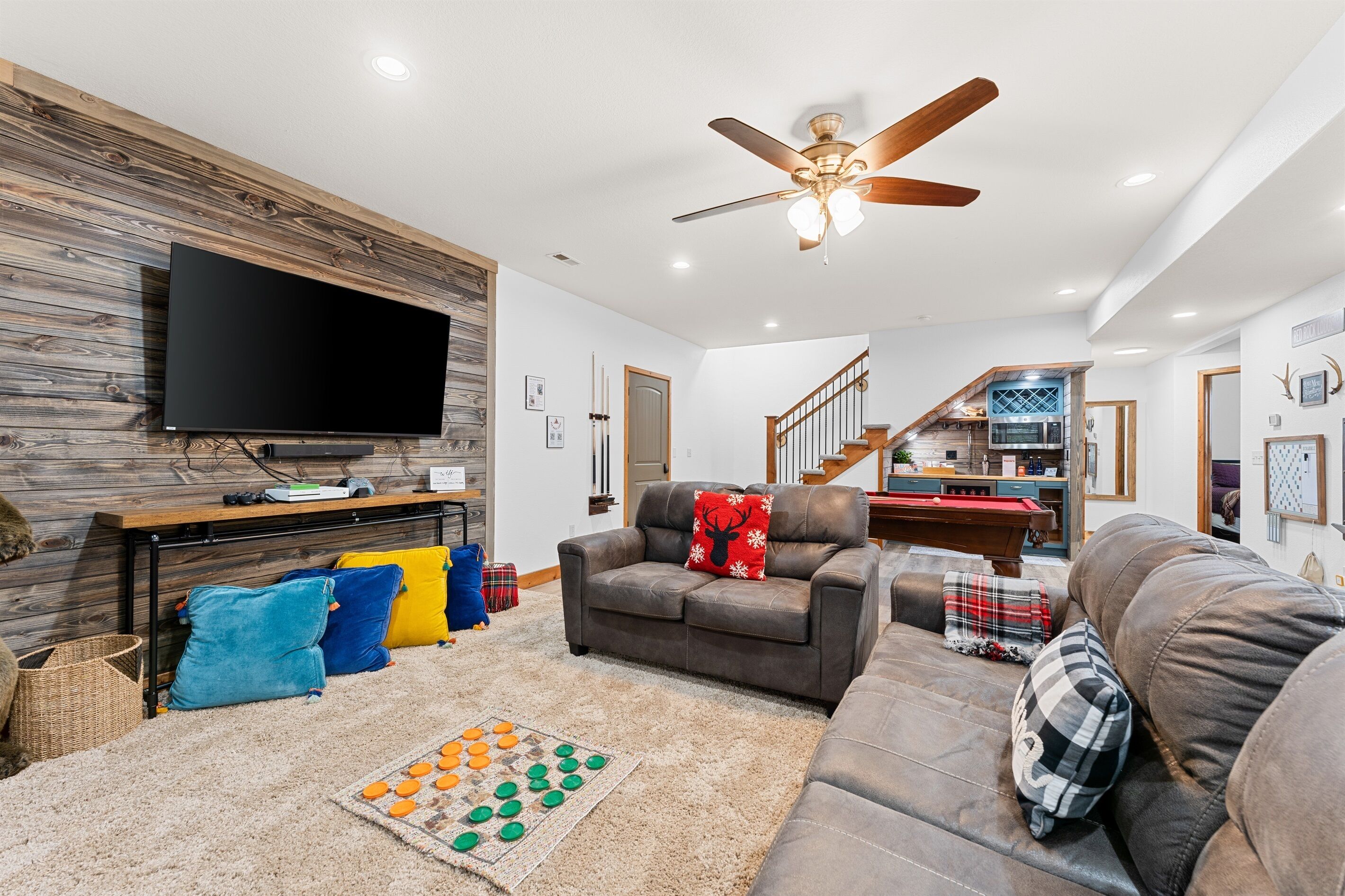 A spacious living room with a flat-screen TV mounted on a wooden wall, a ceiling fan, and a staircase leading to an upper level.