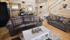 Ein gemütliches Blockhauss-Interieur mit einer Sofagarnitur, einem Esstisch, einer Küchenzeile und einer Treppe.