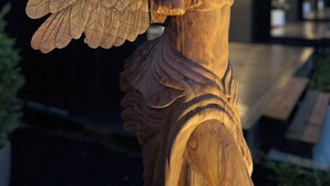 Une sculpture en bois représentant un ange aux ailes déployées, placée devant la façade d’un bâtiment moderne.