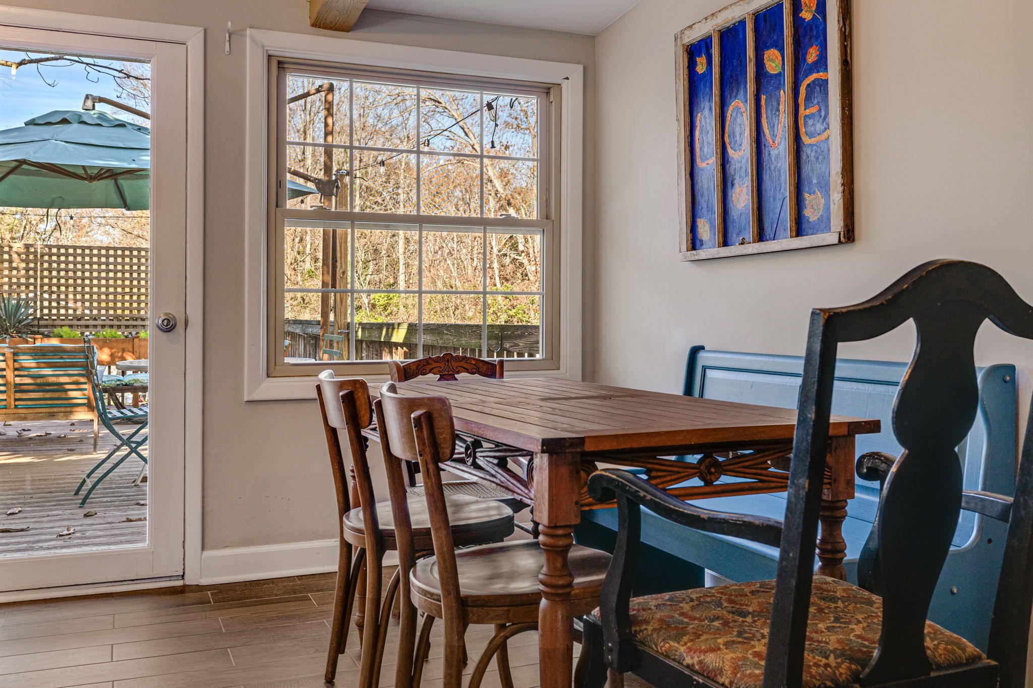Un coin repas avec une table et des chaises en bois, une grande fenêtre donnant sur l’extérieur et un mur bleu orné d’un panneau indiquant le mot « AMOUR ».