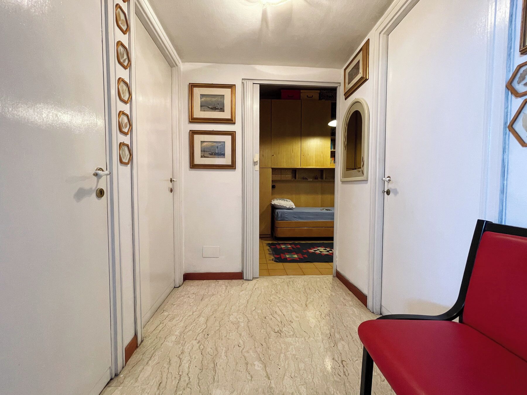 Un pasillo estrecho con una silla roja, una cama con colchón azul y armarios de madera.