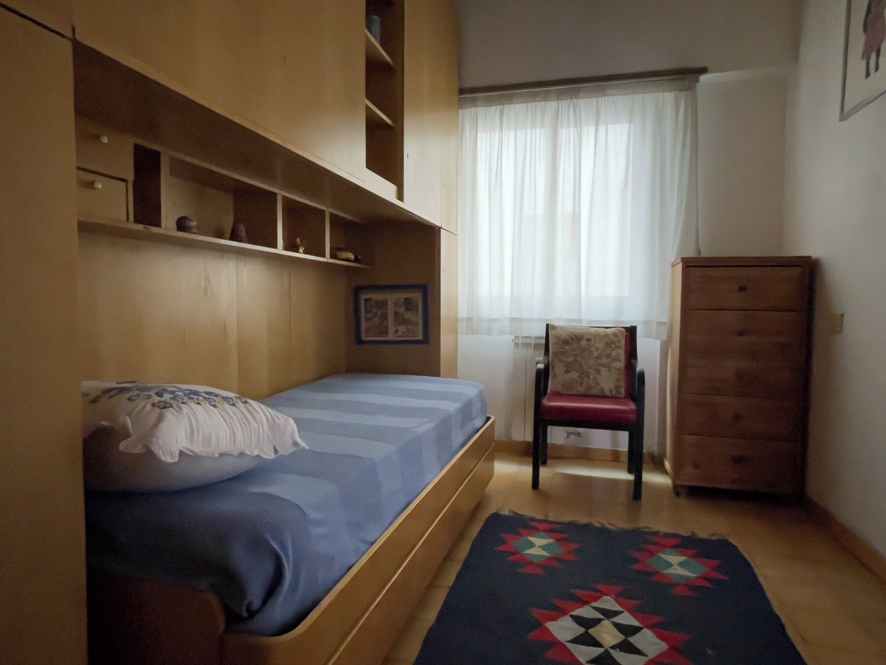 Une petite chambre à coucher, chaleureuse et accueillante, avec un lit simple, une chaise, une commode et une fenêtre ornée de rideaux légers.