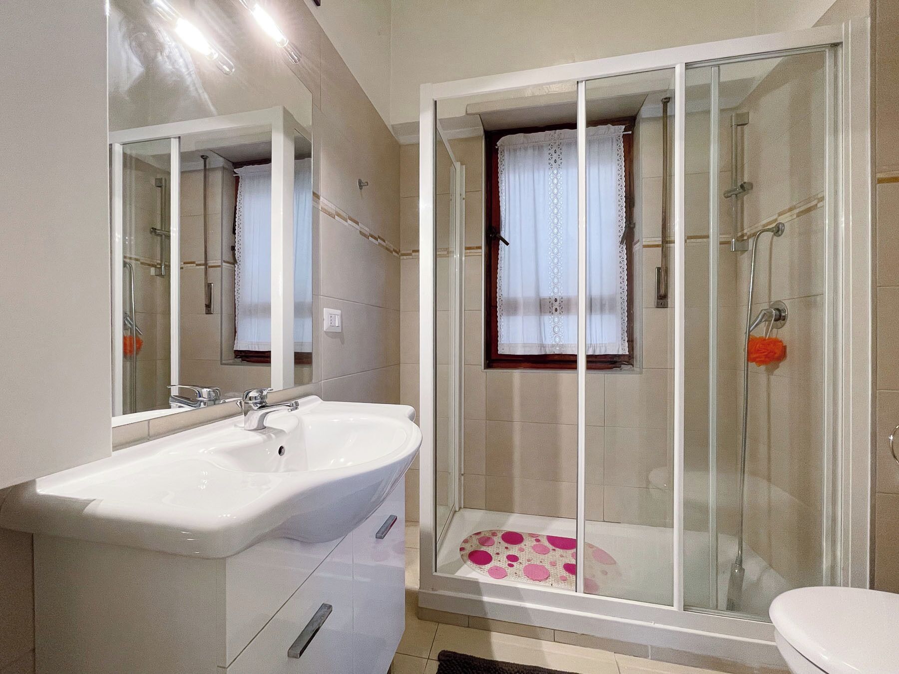A bathroom with a white sink, a glass shower enclosure, and a window with lace curtains.