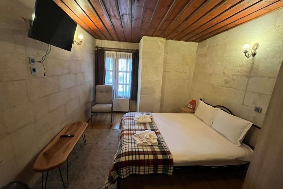 A small, cozy room with a bed, a bench, a chair, a TV mounted on the wall, and a window with curtains.