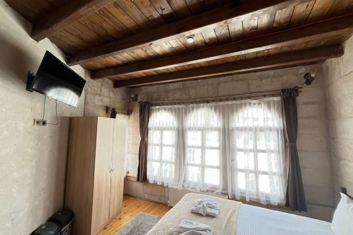 A bedroom with a wooden ceiling, a bed with a white bedspread, a television mounted on the wall, and a window with curtains.
