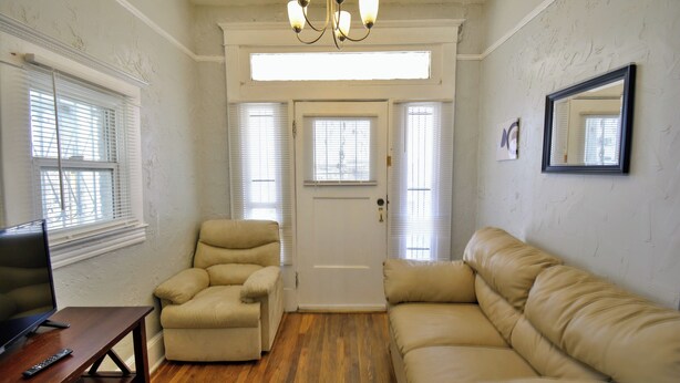 A room with a wooden floor, beige leather sofa, a leather armchair, a wooden table with a remote control, and a door with a window. - Entire Home Near UTEP  (El Paso)