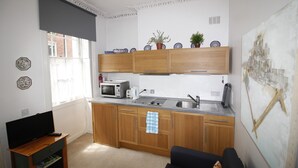 A compact kitchen with wooden cabinets, a microwave, and a sink. There is a television on a stand and a painting on the wall.