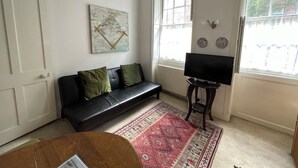 A living room with a black leather sofa, a red patterned rug, a television on a stand, and a large window with a view of the outside.