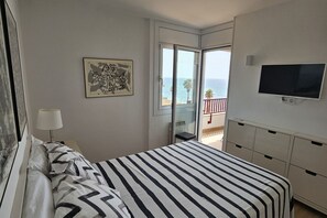 A bedroom with a bed, a TV, a painting, and a balcony with a view of the sea.