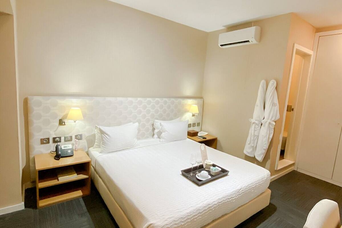 A hotel room with a large bed, bedside tables with lamps, an air conditioning unit, and a pair of white robes hanging on the wall.