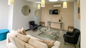 Ett modernt vardagsrum med en beige soffa, ett soffbord och två fåtöljer. En platt-TV är monterad på väggen, och hänglampor hänger från taket.