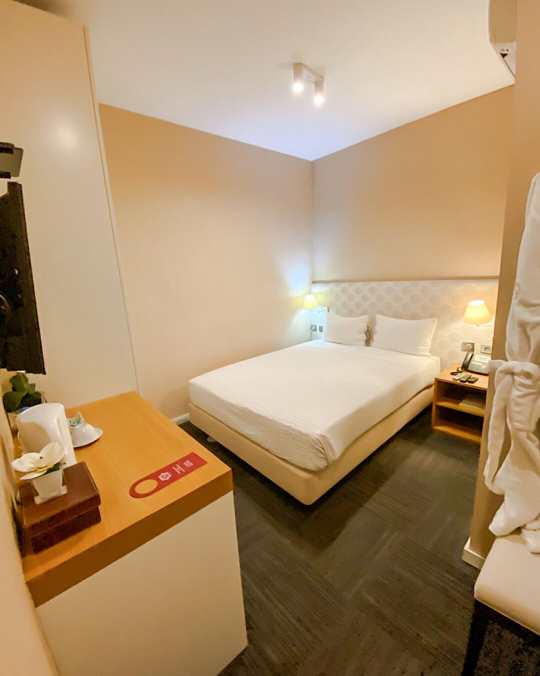 Habitación de hotel con cama, mesita de noche, lámpara y un pequeño escritorio con teléfono y planta.